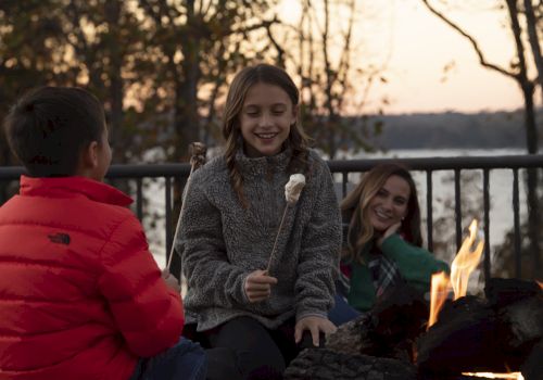 Three people gathered around a campfire, toasting marshmallows and enjoying the evening outdoors by a lakeside with trees in the background.