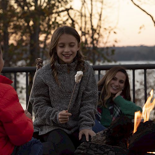 Three people gathered around a campfire, toasting marshmallows and enjoying the evening outdoors by a lakeside with trees in the background.