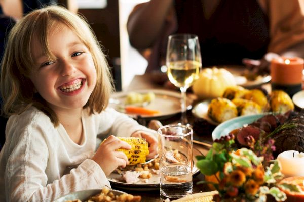 A smiling child is enjoying corn on the cob at a festive dinner table with various dishes, flowers, and candles, creating a warm, celebratory atmosphere.