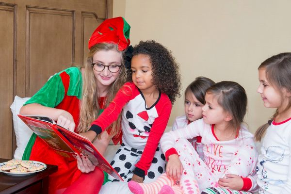 A person dressed as an elf reads a Christmas story to a group of children in pajamas. A plate of cookies and a glass of milk are nearby on a table.