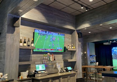 The image shows a bar area with a TV displaying a sports game, a counter with bottles and taps, and a golfing simulator in the background.