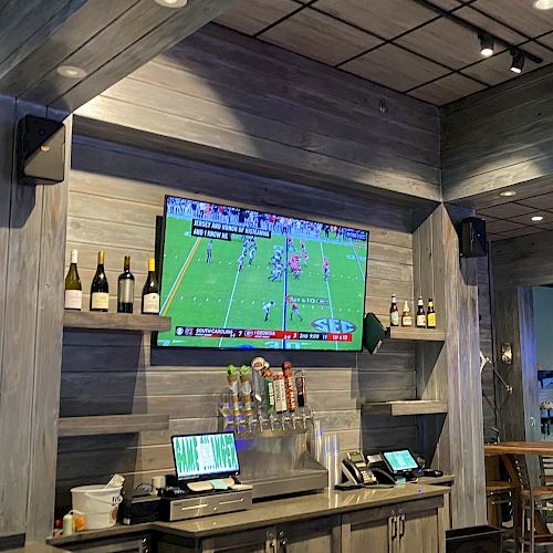 The image shows a sports bar with a TV displaying a football game, several drink taps, bottles of wine, and a golf simulation area in the background.