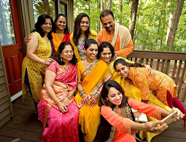 A group of people dressed in traditional attire are smiling and posing for a photo on a wooden deck, surrounded by trees in the background.