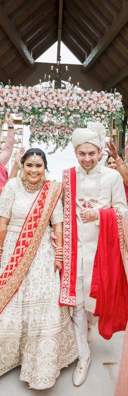 A bride and groom in traditional attire walk through a lively arch of jubilant guests in pink outfits, raising their arms in celebration.