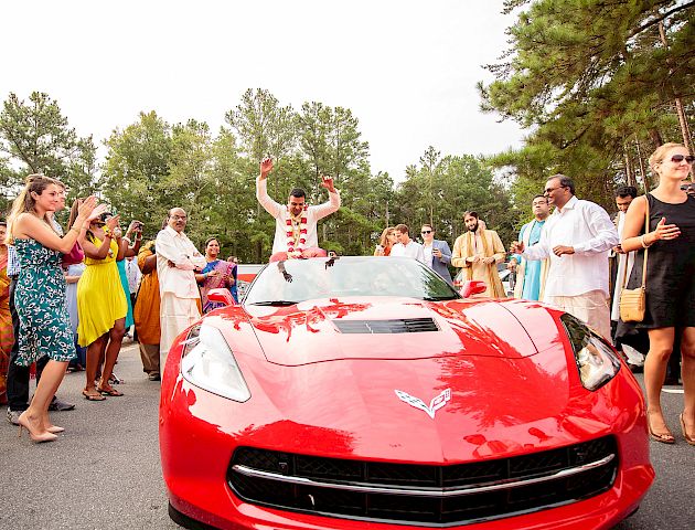 A joyful celebration surrounds a red sports car with a person standing and cheering out of the sunroof, surrounded by people in festive attire.