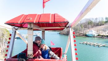 A couple is sitting in a red ferris wheel gondola, overlooking a scenic body of water and docks.