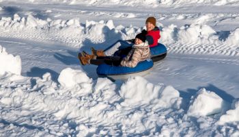 Two people are snow tubing, sitting on inflatable tubes, sliding down a snowy slope. They are dressed in winter clothing.