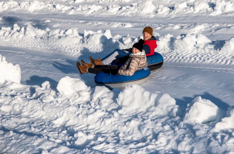 Two people are snow tubing, sitting on inflatable tubes, sliding down a snowy slope. They are dressed in winter clothing.