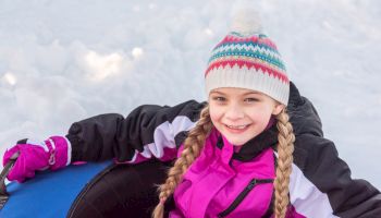 A young girl with braided hair and a knit hat is smiling while sitting on a snow tube in a snowy environment.