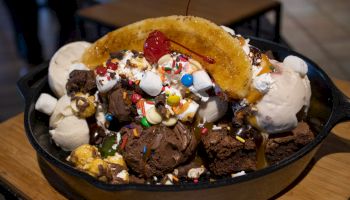 A skillet filled with ice cream scoops, brownies, a banana, whipped cream, marshmallows, sprinkles, M&Ms, caramel, and a cherry on top.