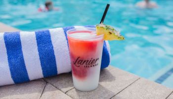 Tropical drink with a pineapple slice, blue and white towel by a pool with swimmers. Container text reads 