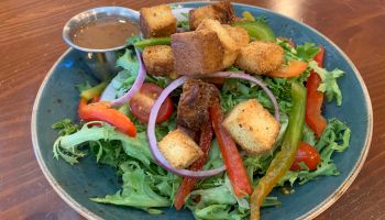 A fresh salad with croutons, red onion rings, bell peppers, and various greens next to a small container of dressing on a wooden table.