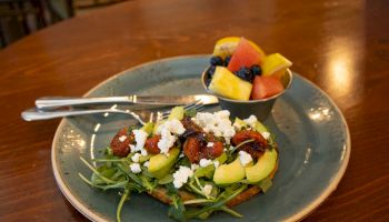 A plate with avocado toast topped with arugula, feta cheese, and sundried tomatoes, alongside a small bowl of mixed fruit.