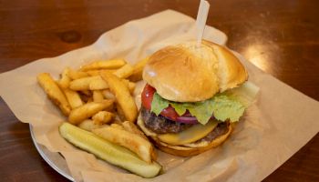 A burger with cheese, lettuce, tomato, and red onion, served with a side of fries and a pickle spear on a brown paper-lined plate.