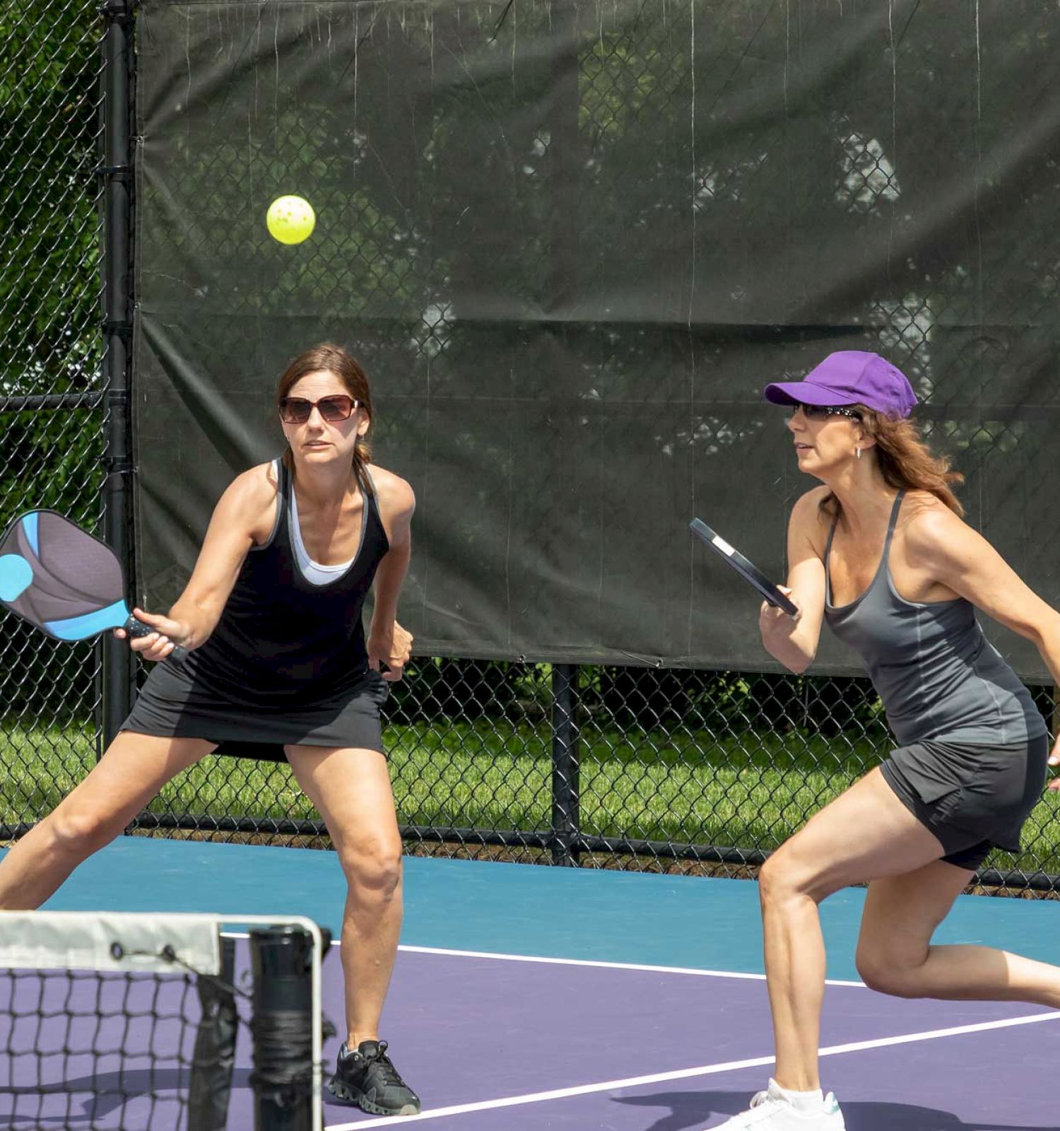 Two women are playing pickleball on an outdoor court; one is about to hit the ball with a paddle.