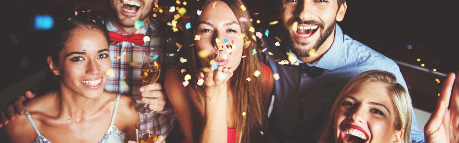A group of five people smiling and celebrating at a party, holding drinks and surrounded by confetti.