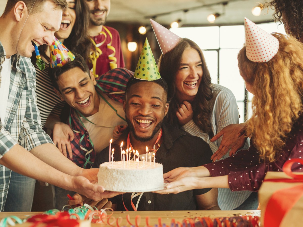 A group of people wearing party hats celebrate a birthday, smiling around a man with a cake topped with candles, in a festive setting.