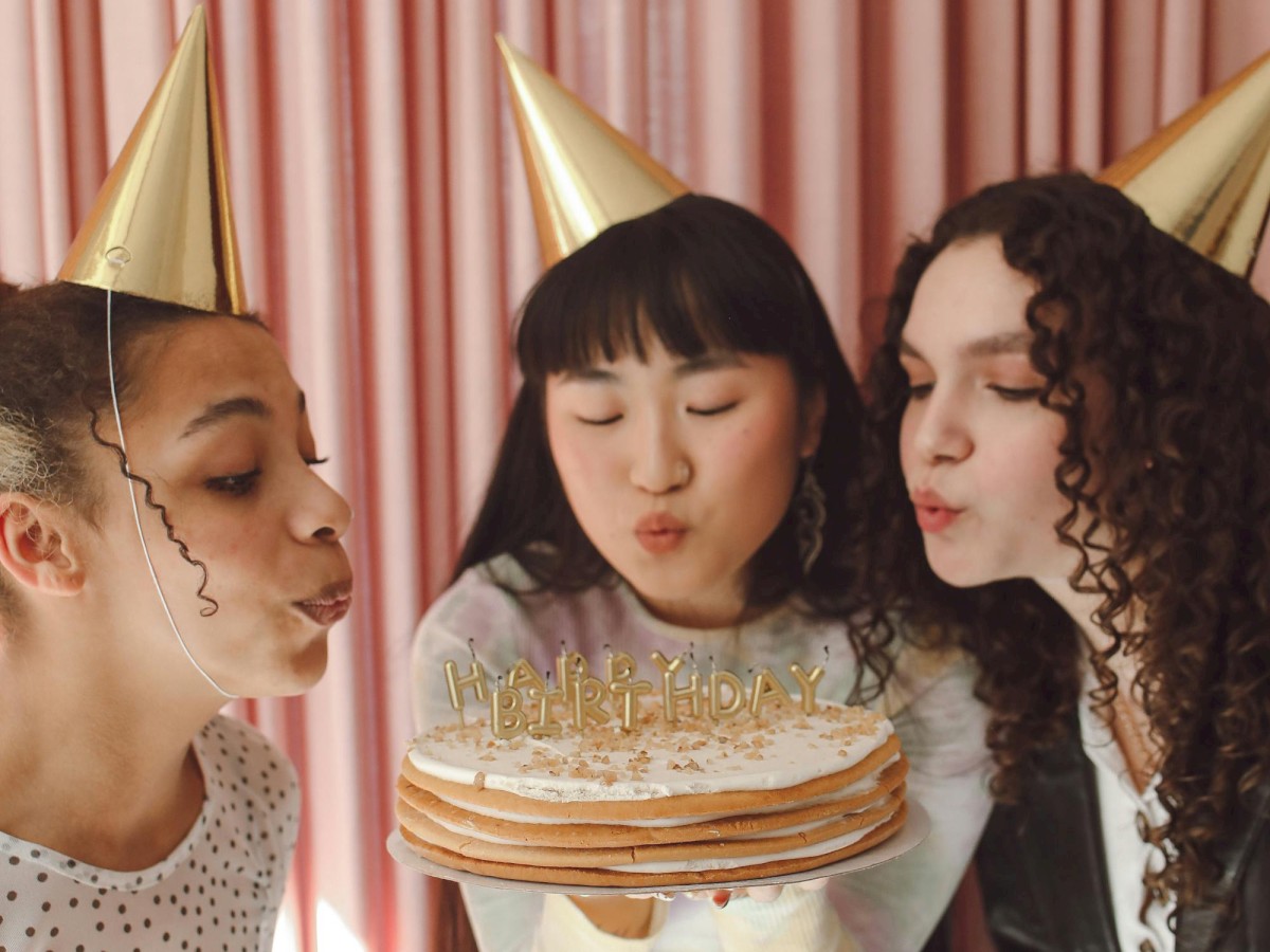Three people wearing party hats are gathered around a cake, appearing to blow out candles. Two are holding cups, and they are in front of pink curtains.