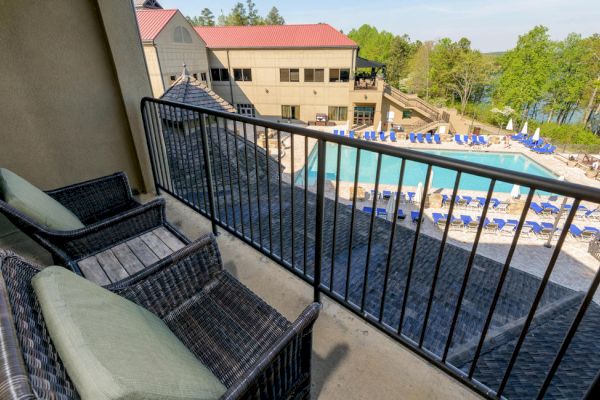 The image shows a balcony with two wicker chairs overlooking a pool area with blue lounge chairs and a nearby building surrounded by trees.