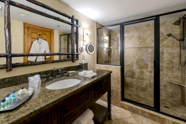 A modern bathroom with a large mirror, vanity counter, toiletries, a robe, and a glass-enclosed shower area with a rain showerhead and wall tiles.