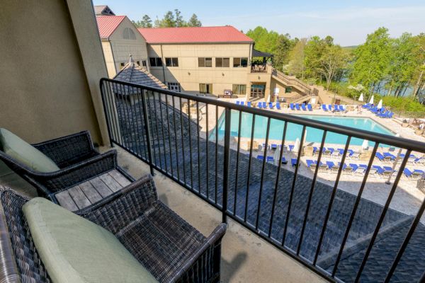 The image shows a balcony with wicker chairs overlooking an outdoor pool area with lounge chairs beside a building, set in a lush, wooded area.
