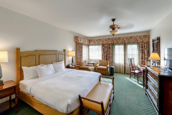 A spacious hotel room with a king-size bed, two nightstands with lamps, a seating area by the windows, a TV, and classic wooden furniture.