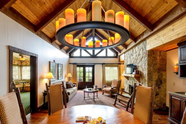 This image shows a cozy living and dining room area with a high wooden ceiling, large modern chandelier, stone fireplace, and rustic furnishings.