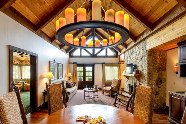 The image shows a cozy living area with wooden interiors, a round chandelier, a stone fireplace, and a dining table set up, creating a warm atmosphere.