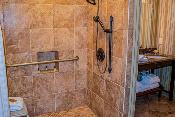 A tiled shower with a grab bar, showerhead, and niche, adjacent to a vanity with towels and toiletries on a wooden shelf.