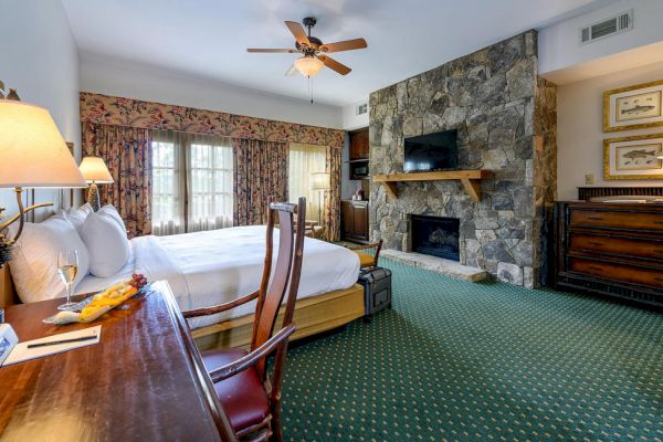 This image shows a cozy hotel room with a large bed, stone fireplace, desk with a lamp, and a TV. The decor includes patterned curtains and green carpet.