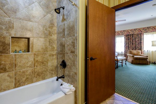 The image shows a bathroom with a tiled shower and a view of a hotel room featuring a bed, armchair, lamp, and windows with curtains.