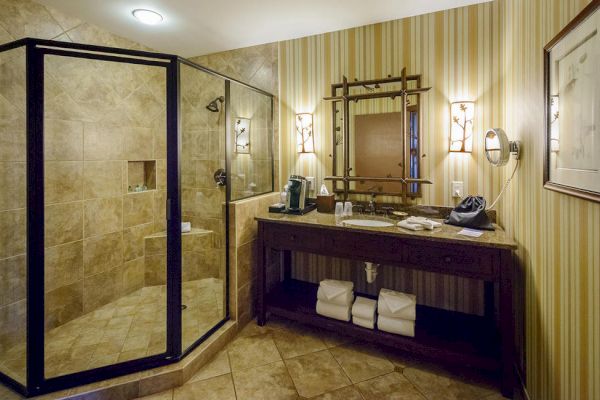 A well-lit bathroom with a glass shower, a wooden vanity with a framed mirror, wall sconces, and striped wallpaper, featuring neatly folded towels underneath.