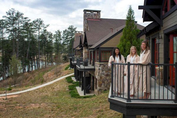 Three people in robes stand on a balcony overlooking a scenic wooded area with a path and a lake, near a rustic-style building.