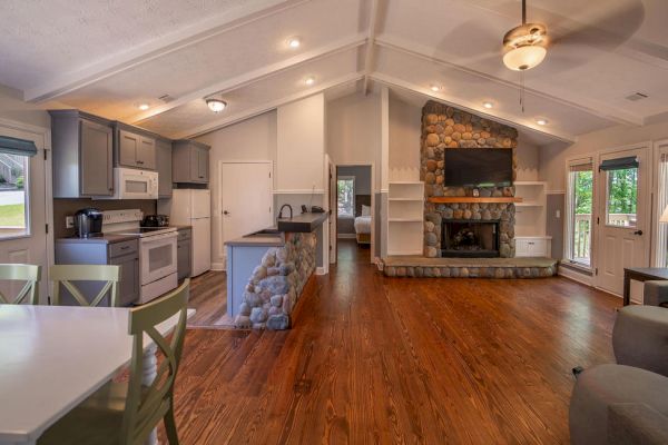 The image shows a cozy open-concept living space with a kitchen, dining area, stone fireplace, and wooden floors.