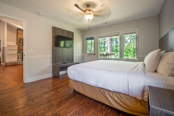 The image shows a bedroom with a large bed, a wall-mounted TV, a ceiling fan, and a view of a wooded area through three large windows.