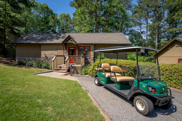 A small cottage with a wooden exterior and a green golf cart park in front under a canopy of tall trees, surrounded by lush greenery and a tidy yard.