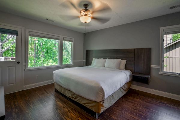 A modern bedroom with a double bed, wooden flooring, ceiling fan, and large windows providing natural light and a view of the outdoors ends the sentence.