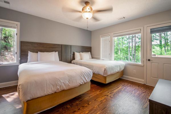 A bedroom with two beds, white bedding, wooden floor, ceiling fan, windows, and a door, all facing an outside view of trees and greenery.