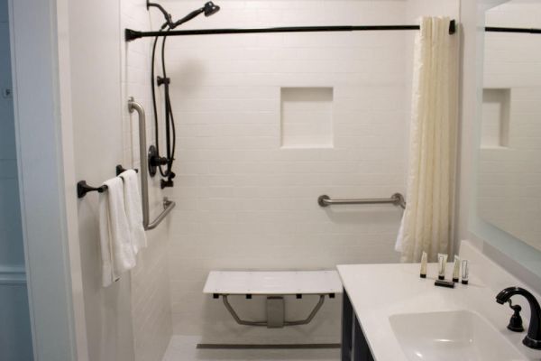 A bathroom with a walk-in shower, a foldable bench, grab bars, a wall-mounted showerhead, a towel rack with towels, and a sink with a mirror.