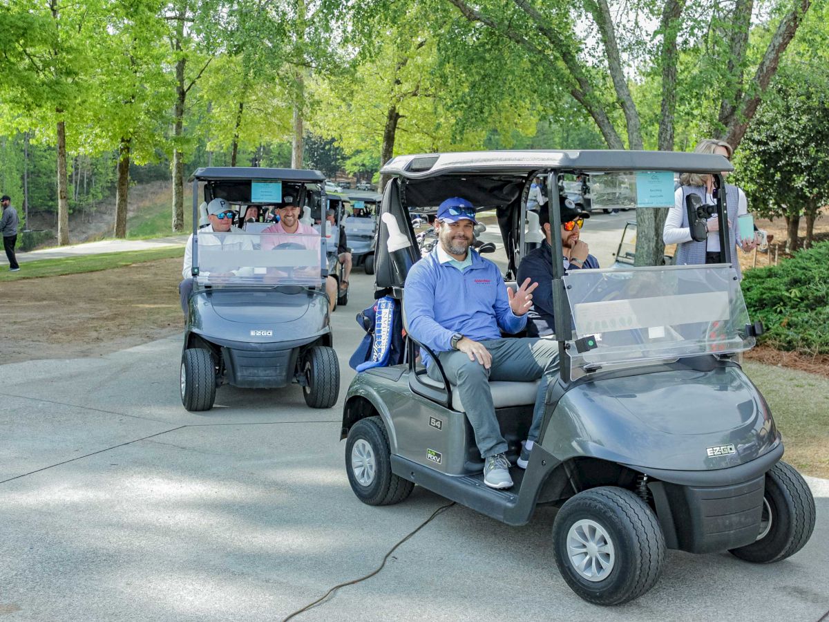People are sitting in golf carts on a paved path, surrounded by trees in a park-like setting. One person in the front cart is waving.