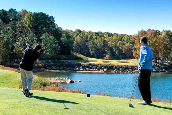 Two people are playing golf by a scenic lake, with a lush forest in the background and one person swinging a golf club.
