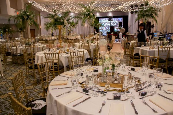 This image shows an elegant banquet hall setup with round tables, gold chairs, floral centerpieces, and string lights strung across the ceiling.