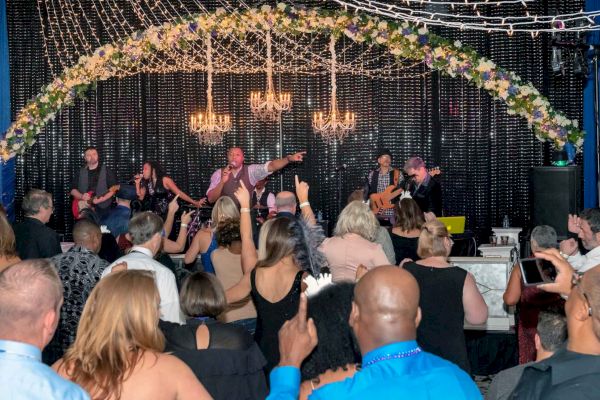 A live band performs on stage under a floral and chandelier arch while a crowd of people in formal attire enjoy the music and dance.