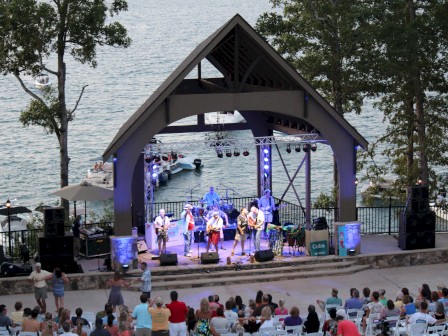 An outdoor concert by a lake with a band playing on stage, surrounded by trees and an audience seated in chairs.