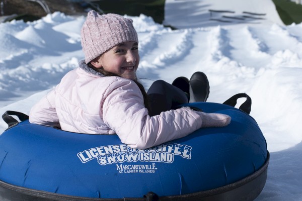 A person in a pink jacket and beanie is sitting on a blue snow tube on a snowy slope, smiling back at the camera.