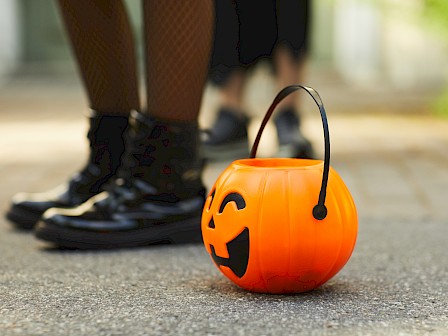 An orange pumpkin-shaped bucket with a handle on the ground, likely for Halloween treats; two people in boots stand behind it.