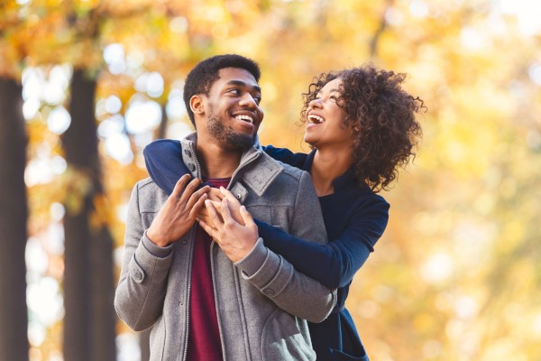 A woman playfully hugs a smiling man from behind in an outdoor setting with autumn foliage in the background.