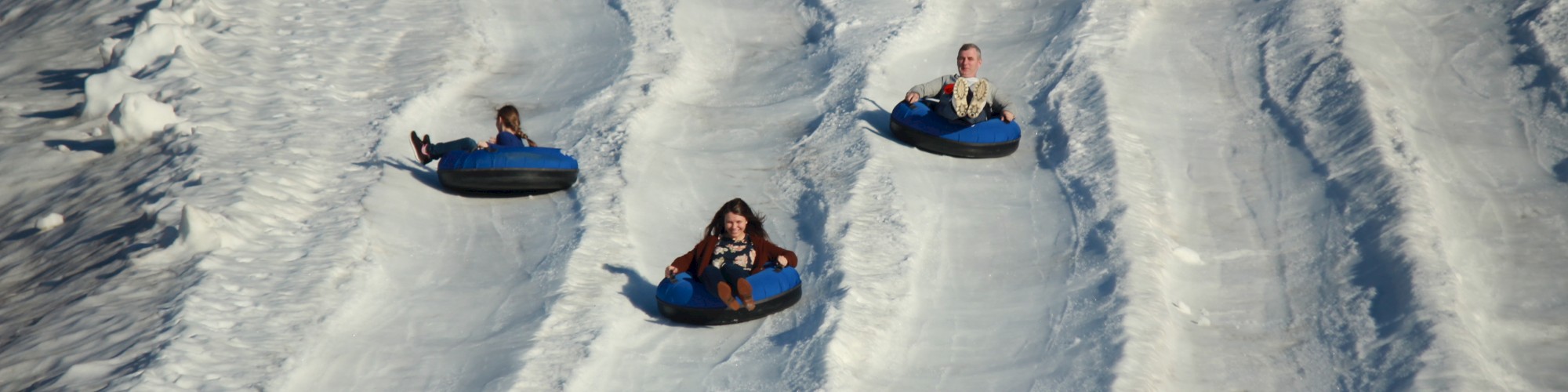 Three people are sliding down snowy lanes on inner tubes in an outdoor winter activity, appearing to race each other down the slopes.