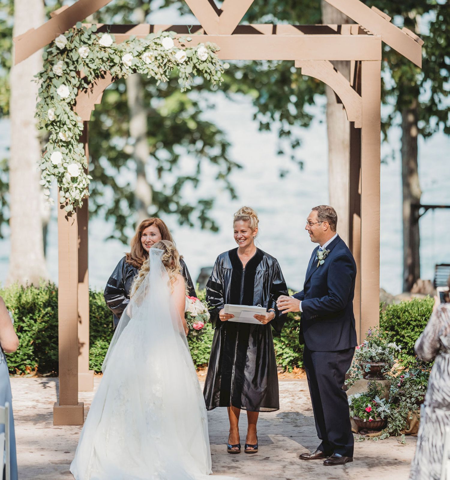 A couple stands under an arch, with an officiant, during an outdoor wedding ceremony. Trees and greenery are visible in the background, ending the sentence.