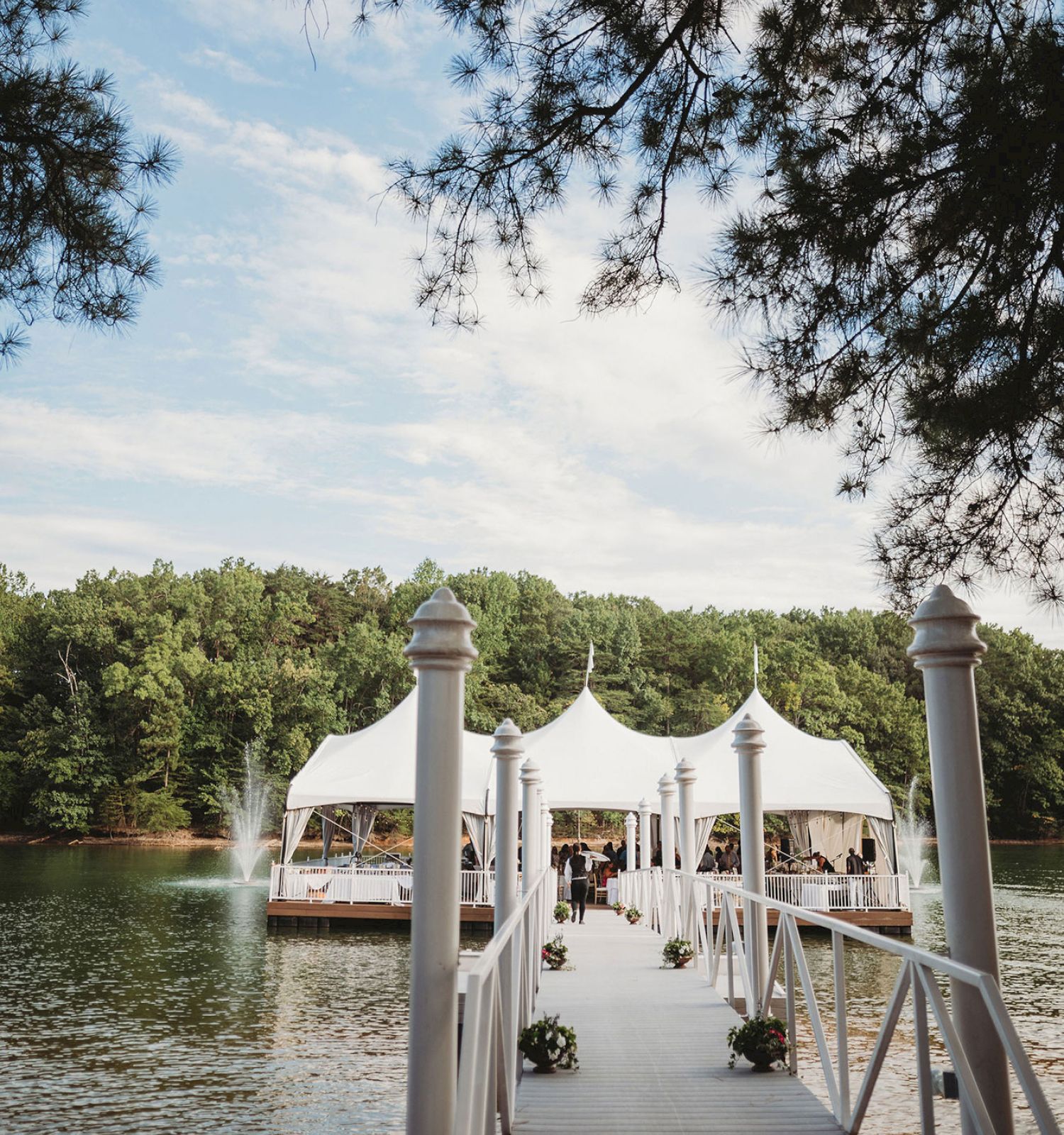 A wooden dock leads to a white tent setup on water, surrounded by trees under a clear sky, with some potted plants along the walkway.